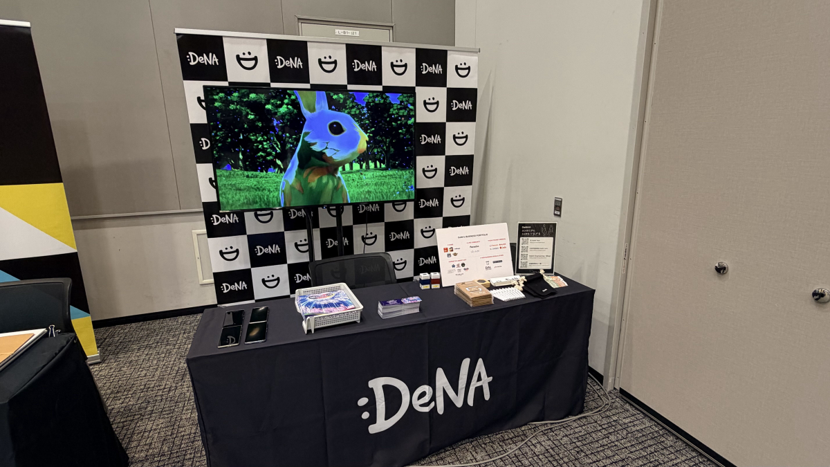 DeNAのロゴが入ったテーブルクロスが掛けられた展示ブース。机の上に資料や展示物が置かれ、背後の市松模様のパネルの前にはキャラクターの映像が流れるモニターが設置されている。