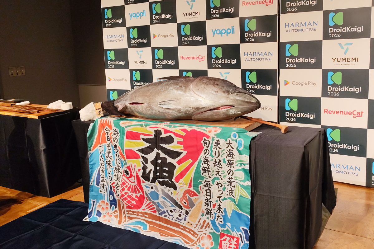 「DroidKaigi 2026」などのスポンサーロゴが並ぶバックパネルを背景に、テーブルの上に横たわる巨大な丸ごとのマグロの写真。テーブルの手前には「大漁」と大きく書かれた色鮮やかな大漁旗が掛けられており、マグロの奥には解体用の長い包丁が置かれている。