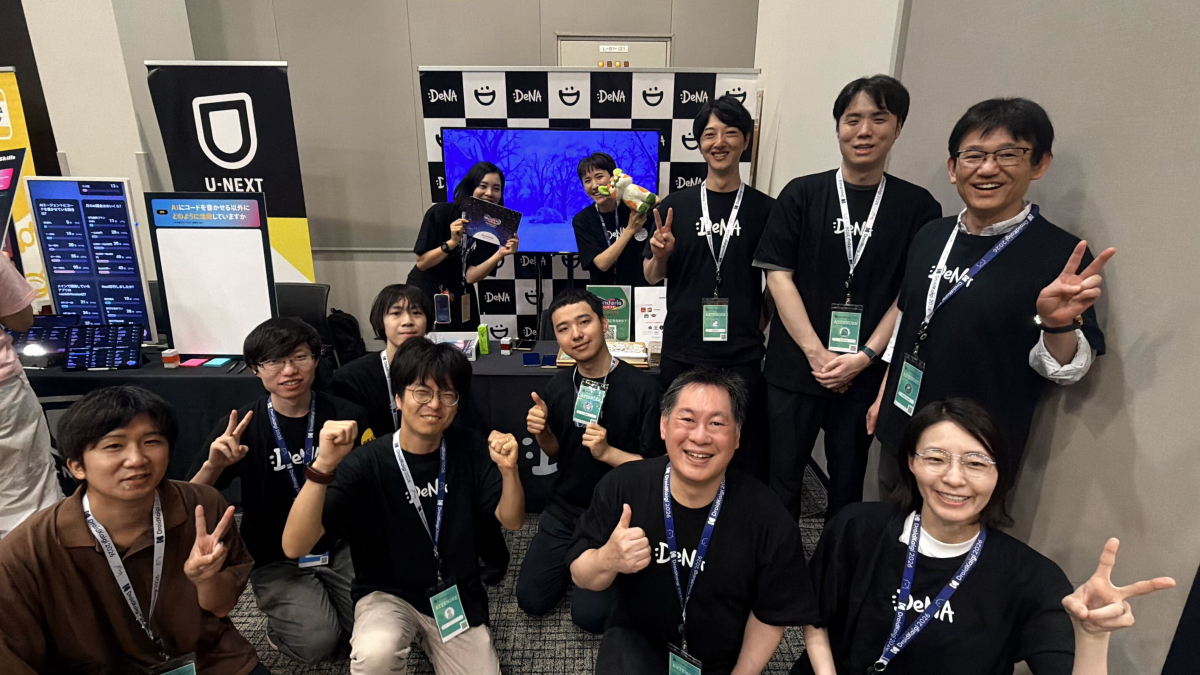 DeNAのイベントブースにて、お揃いの黒いロゴ入りTシャツを着たスタッフたちが笑顔でピースサインやガッツポーズをしている集合写真。